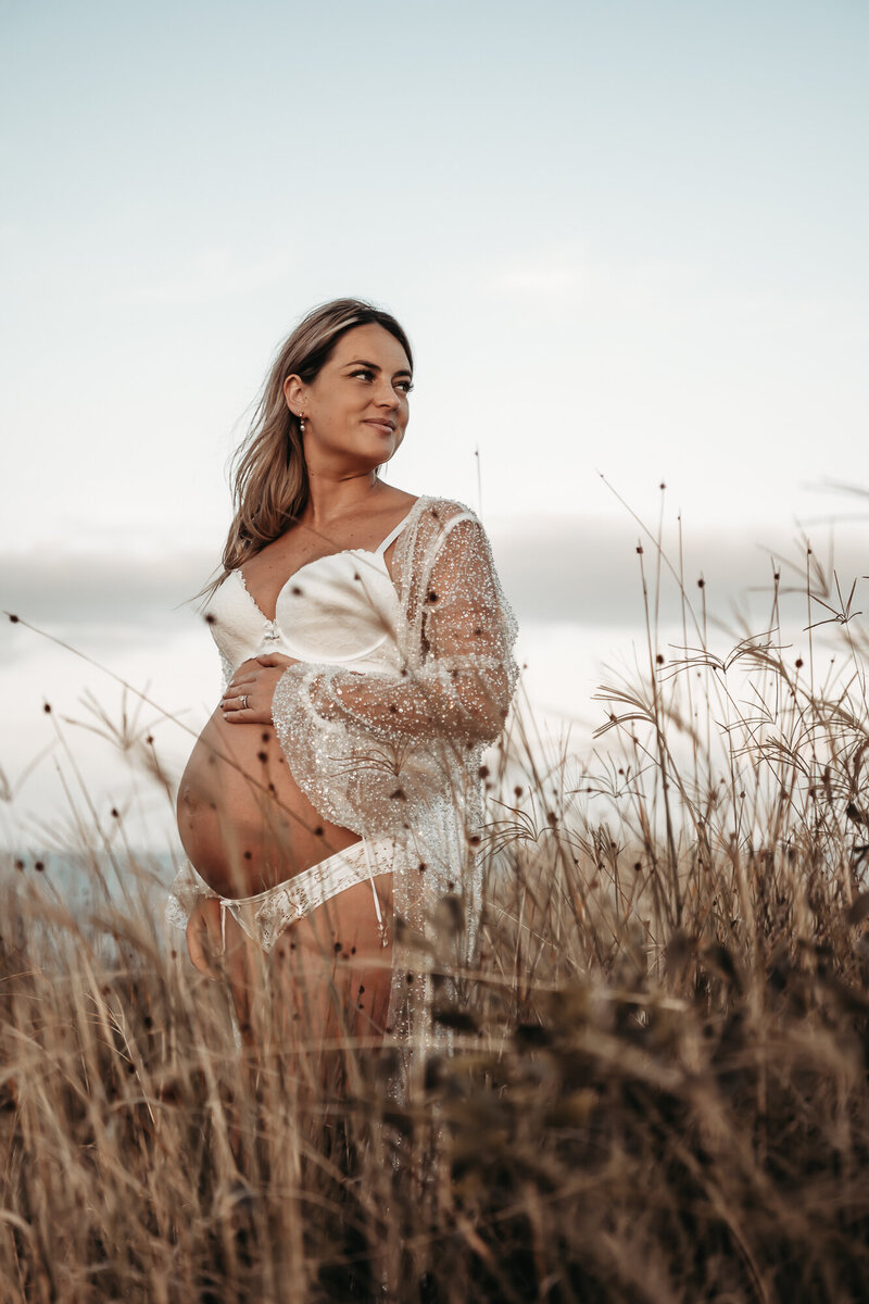 Pregnant woman in floral off-shoulder dress stands in a forest, one hand on her head, the other on her exposed belly—an exquisite moment captured by a Wollongong maternity photographer.