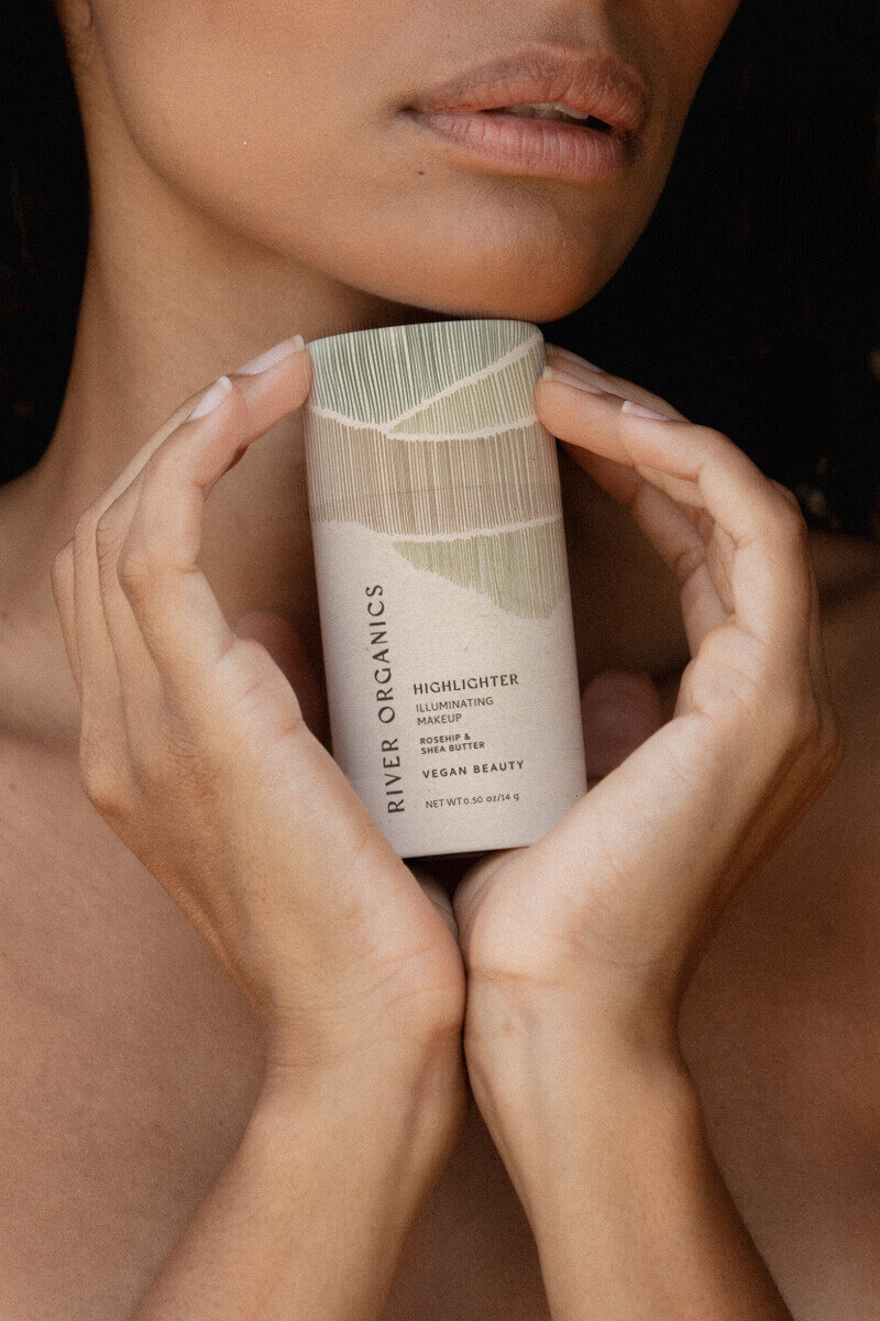 A close-up of a woman holding the River Organics Dusty Rose Blush Stick near her chest, with the product's eco-friendly packaging displayed prominently. The image reflects the brand’s emphasis on natural, clean beauty and sustainability.