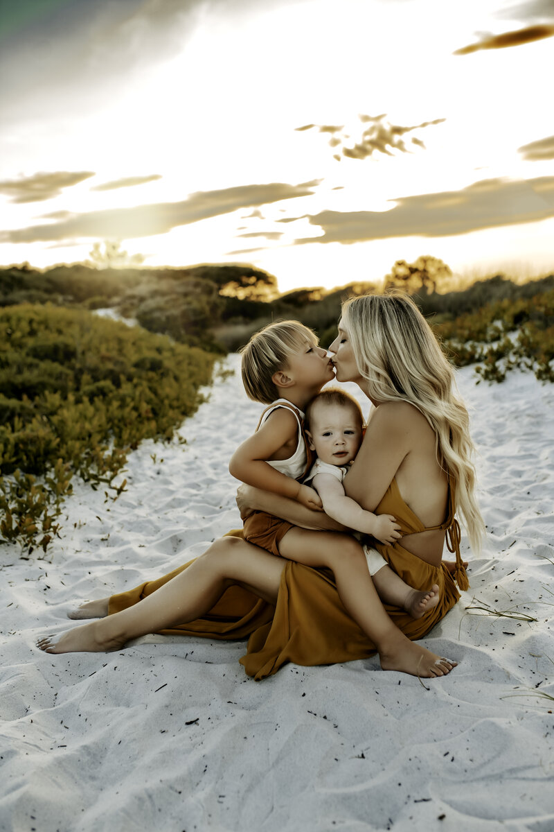 Family Photographer, a mother sits in the Florida sand her baby and young boy both in her lap, she kisses her son