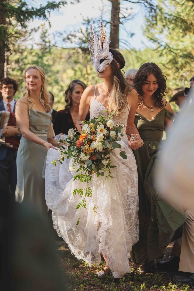 A bride being walked by her bridesmaids to her elopement ceremony in the woods