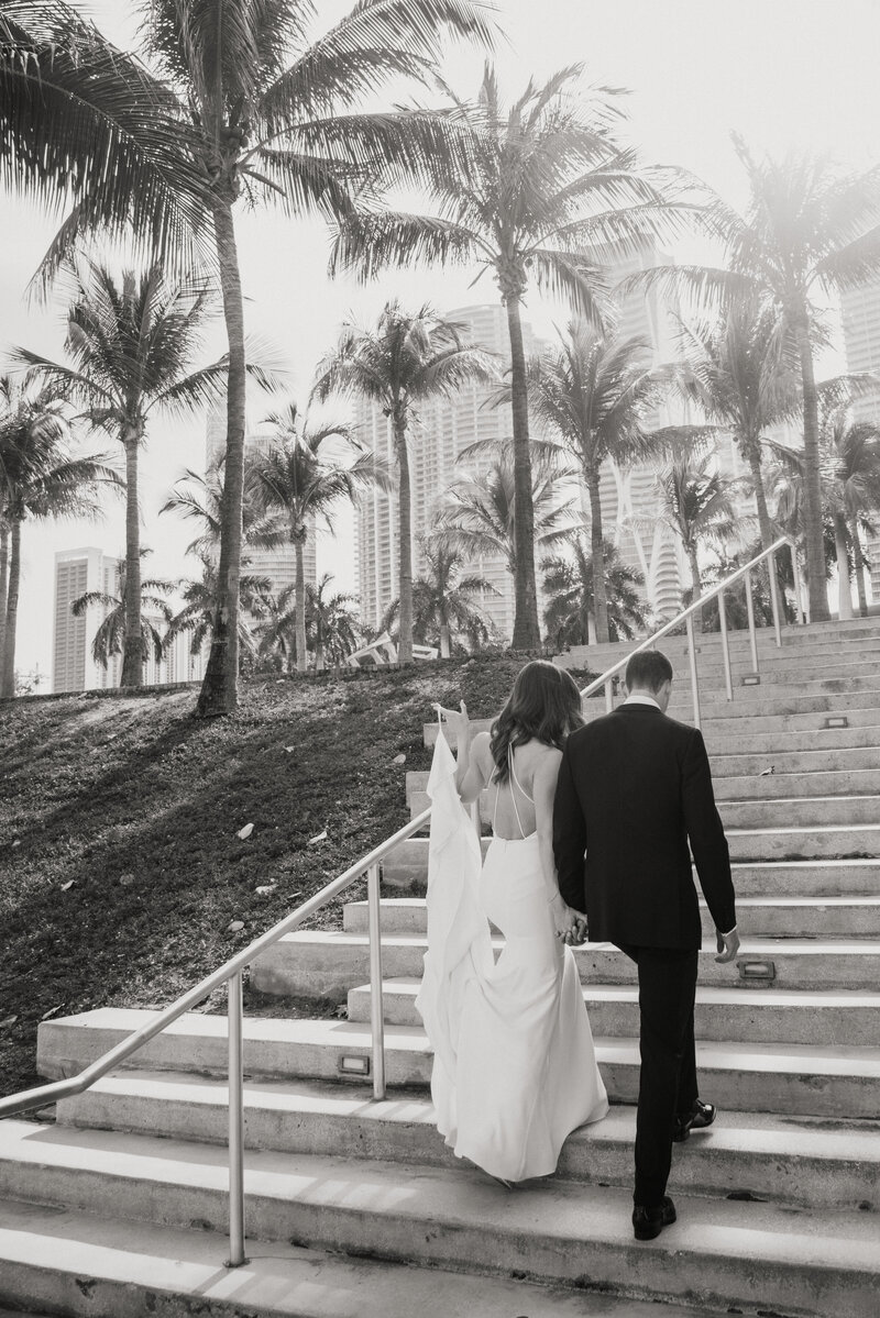 Bride and groom walking up stairs with palm trees and Miami skyline in the background.