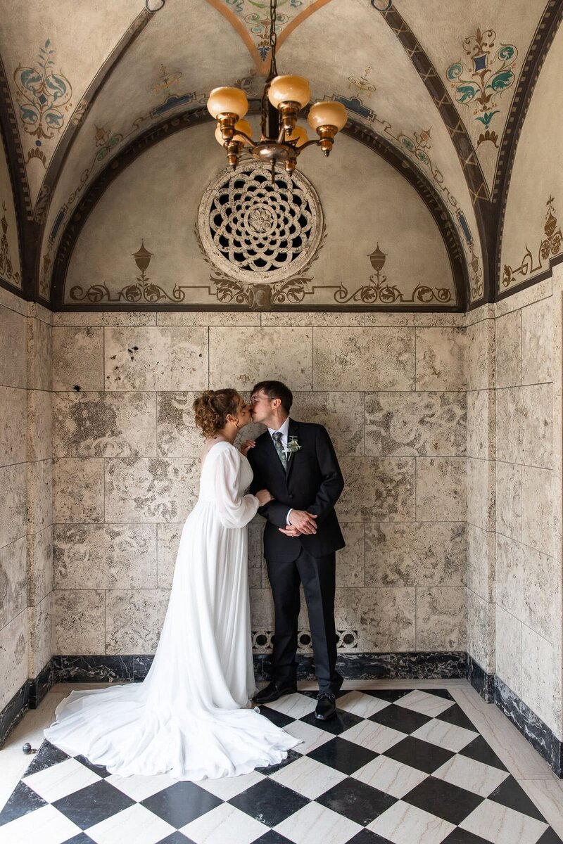 The couple stands inside an arched marble alcove, kissing under a gold vintage chandelier. The bride’s train fans out across the black-and-white tile floor, and detailed floral scrollwork decorates the domed ceiling above them. A romantic, architectural highlight of this unique wedding venue
