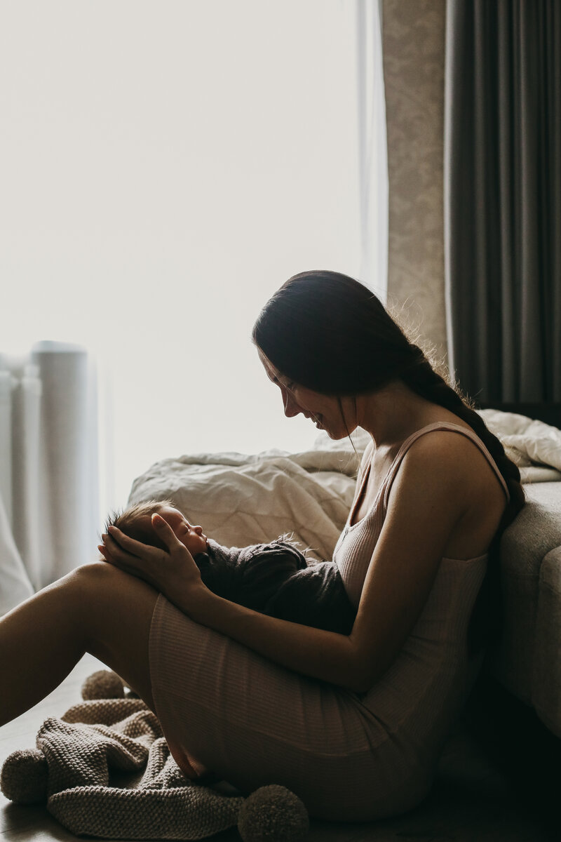 New mom in New Jersey smiling at her newborn baby on her lap, captured in gentle backlit lighting — representing breastfeeding and postpartum support.