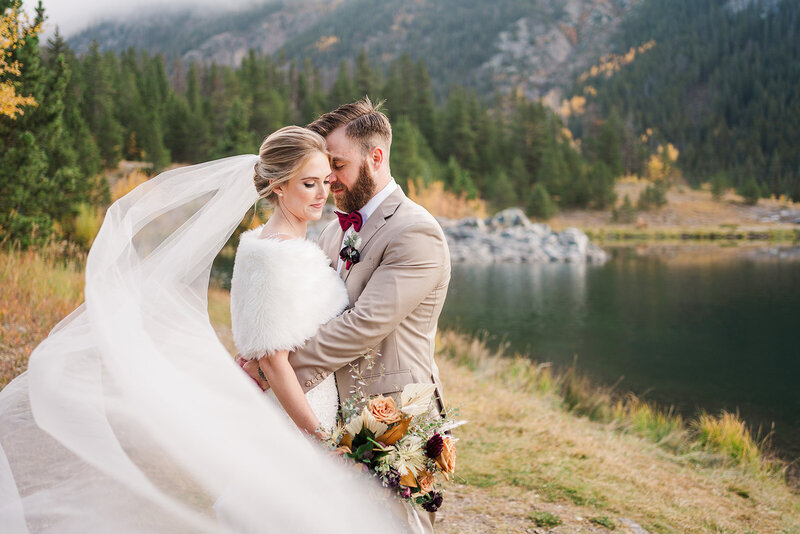 A man holds a woman in a wedding dress in front of a lake and mountains