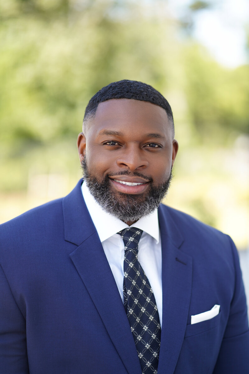 This professional headshot features the Executive Director of the Boys and Girls Club of the Grand Strand in Myrtle Beach, South Carolina. Captured in natural light photography, the subject is posed confidently in a business suit against a clean, neutral background. The soft lighting highlights an approachable yet authoritative presence, reflecting the leadership role in the community.