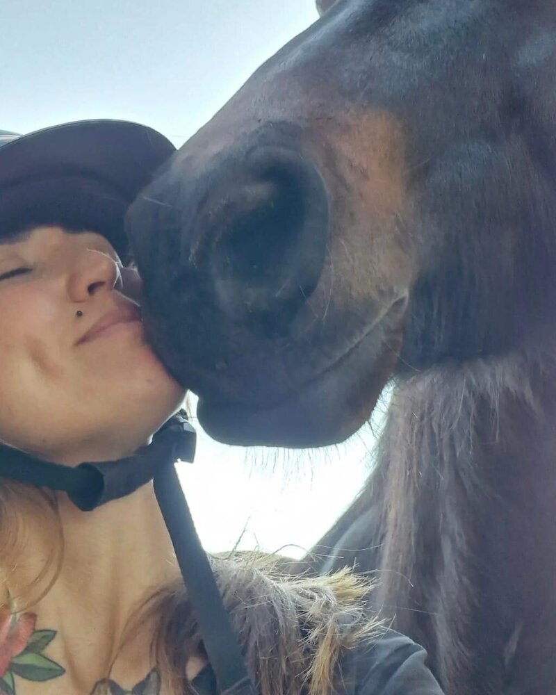 Ashley May (Collet), instructor at Camelot Horse Riding School, smiling while wearing a Camelot-branded vest beside a horse.