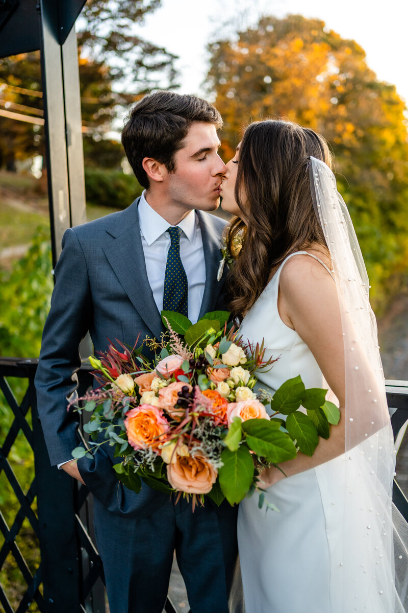 Bride and groom kissing at autumn elopement with colorful bouquet and golden light