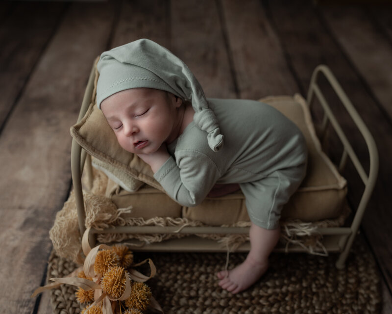 A softly sleeping newborn dressed in sage knitwear on a mini rustic bed styled with vintage textures.