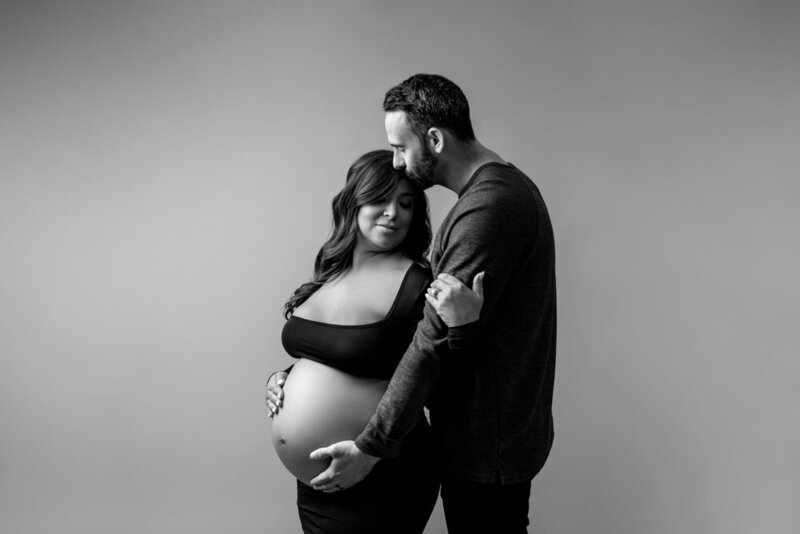 Black and white maternity portrait at Maternity and Newborn Studio of Atlanta with  a pregnant woman in black maternity gown holding her baby bump and embracing her husband.