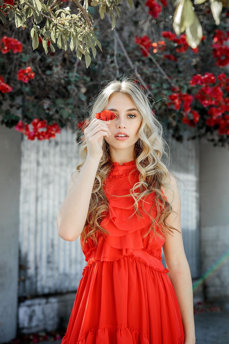 Teen girl in a bold red ruffled dress standing in front of a flowering vine wall, holding a red blossom in front of one eye for a whimsical and fashion-forward pose.