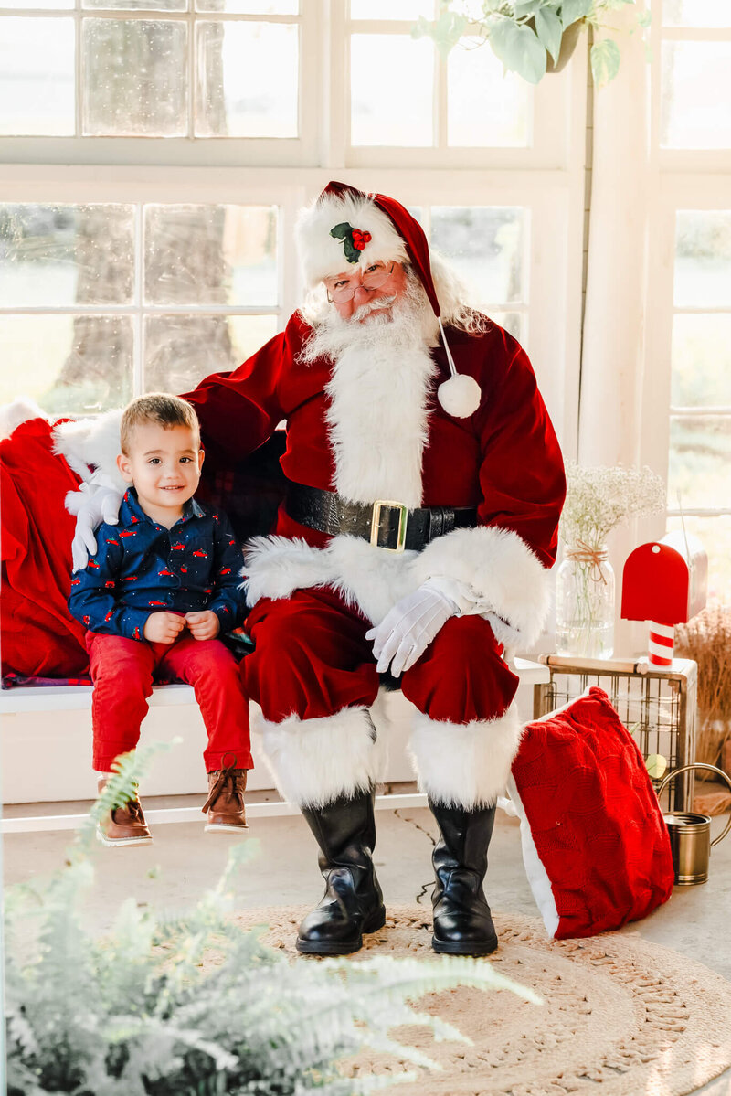 Santa and a little boy in a Christmas outfit sit on a bench in a sun porch. They are both looking at the camera and smiling.