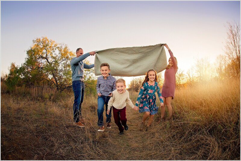 Parents hold a blanket in a field high, and their three children run playfully underneath it.