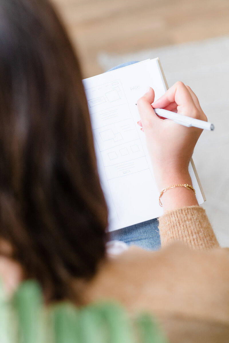 Femme écrivant sur un carnet, un stylo à la main, on aperçoit le schéma d'une page de site Internet