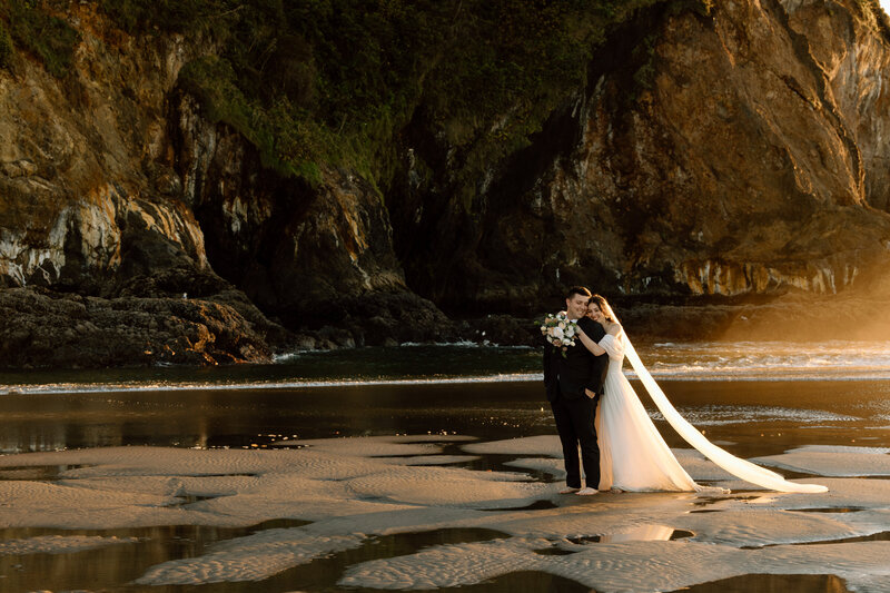 Couple hugging on a beach with a cliff behind them.