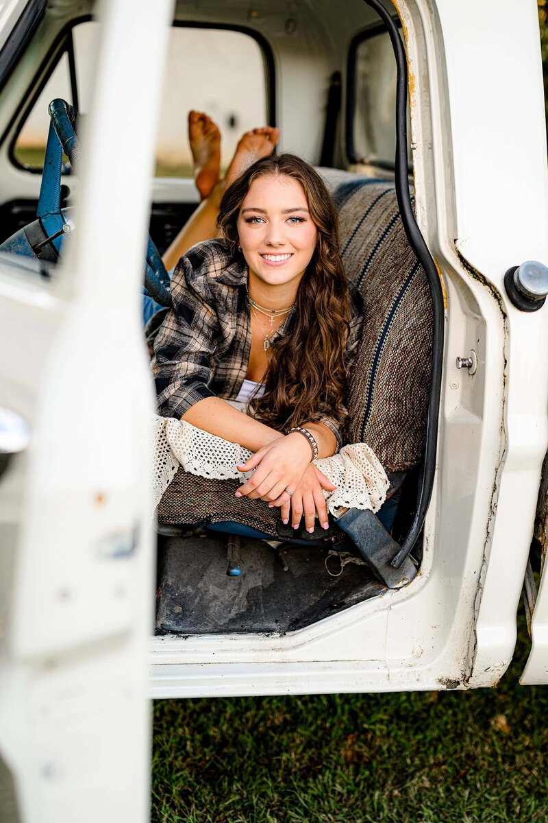 senior girl is laying on her belly in a truck seat wearing a flannel and white shirt