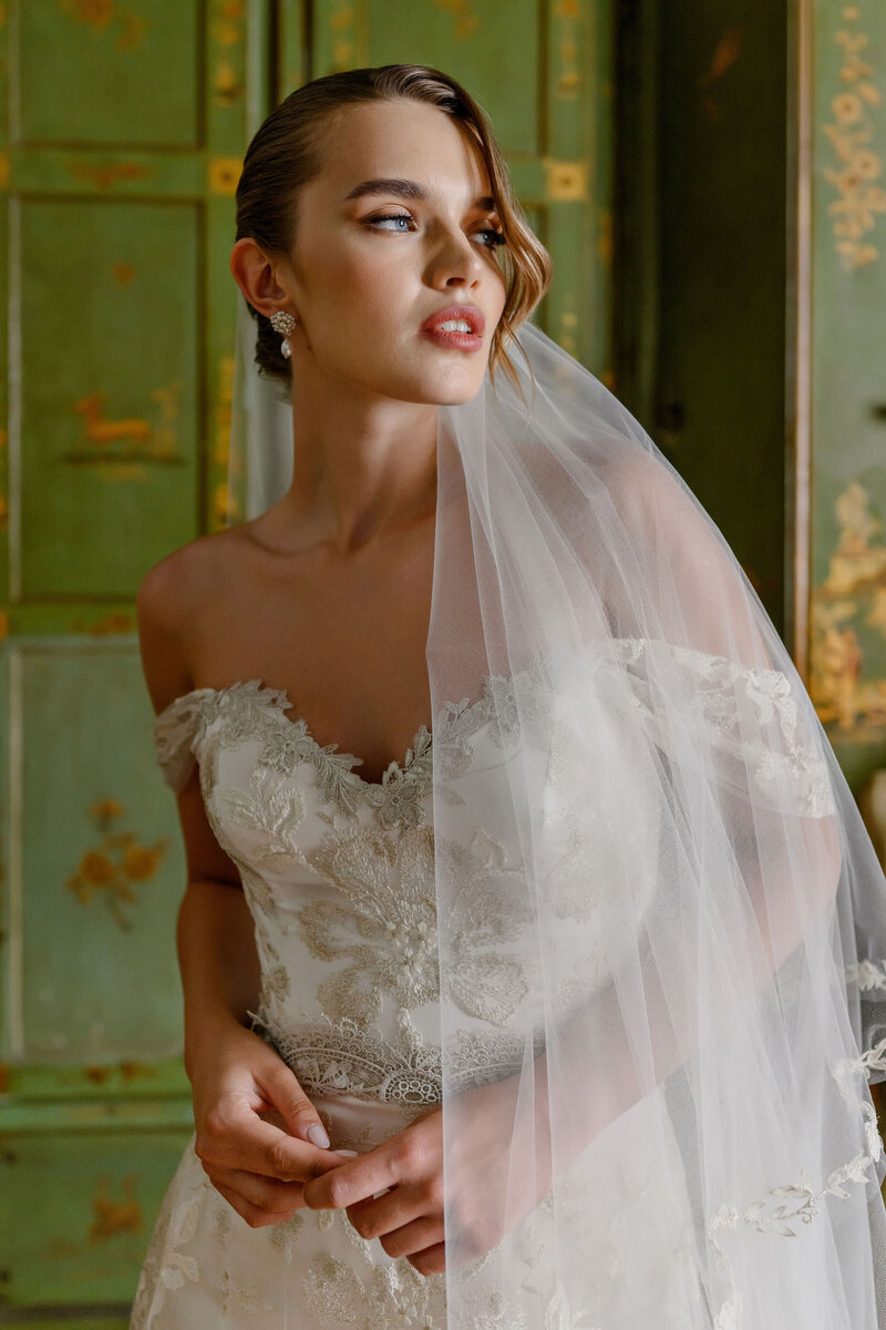 A bride in an intricate lace gown and veil gazes confidently in a beautifully styled room.