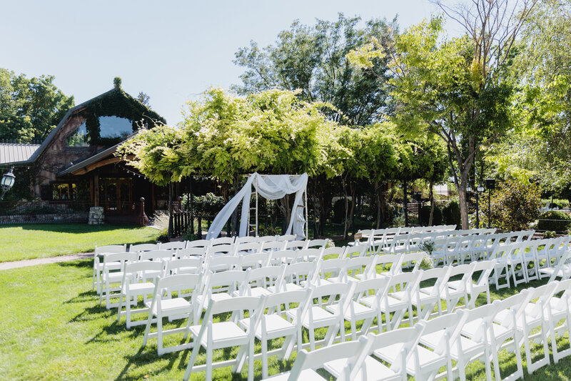 White ceremony chairs are set up in front of a vine wrapped gazebo