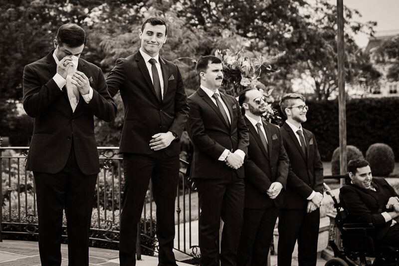 Emotional groom wiping tears as groomsmen look on during outdoor wedding ceremony in New Jersey