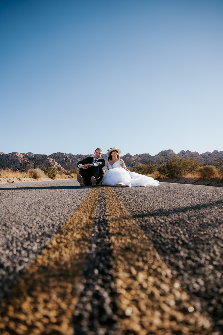 Bride and groom in wedding attire sitting together on a desert highway in Joshua Tree National Park, with a scenic mountain backdrop and clear blue sky. Photo by Auburn Blue Photography.