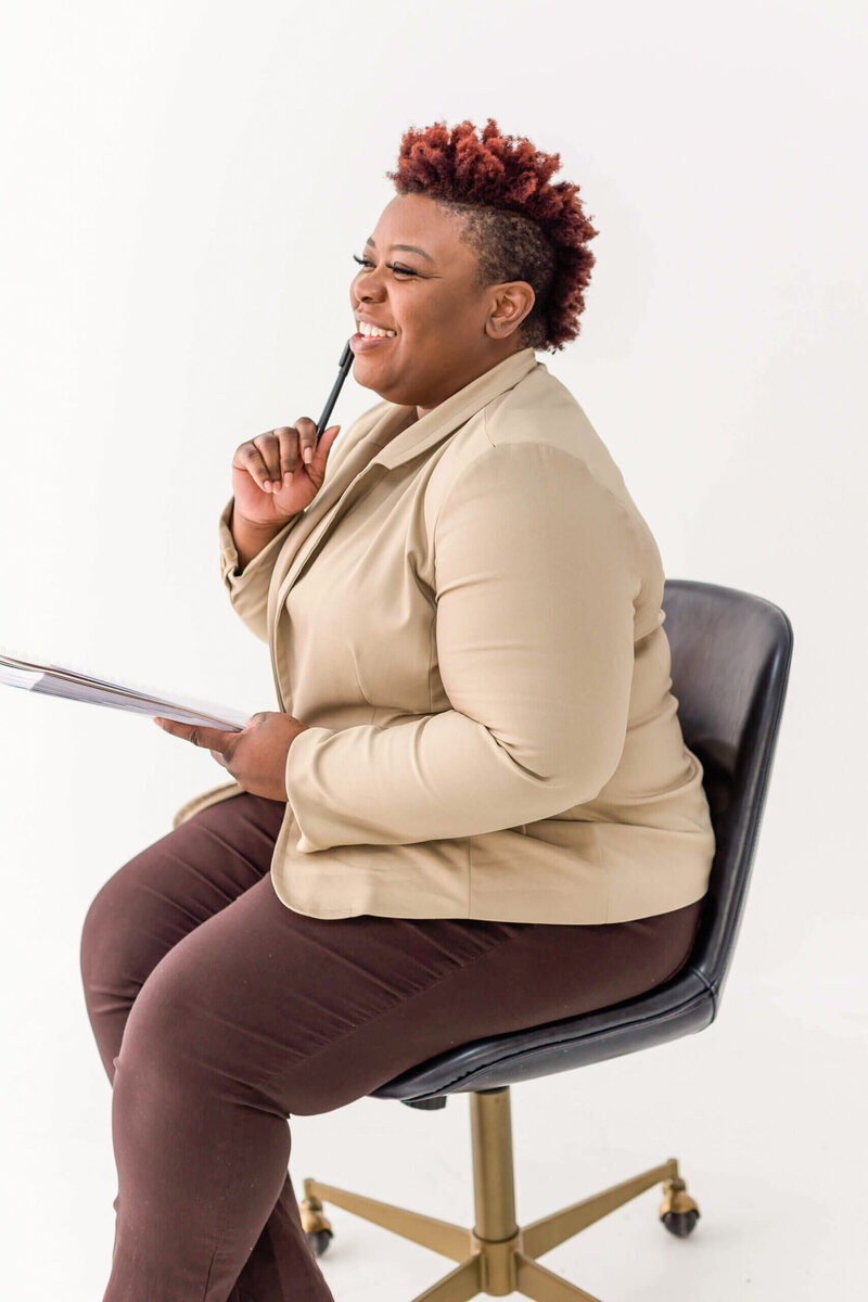A female entrepreneur with short curly hair, dressed in a beige blazer and brown pants, sits confidently on a swivel chair. She holds papers and a pen, smiling as she gazes into the distance, embodying vision and ambition.