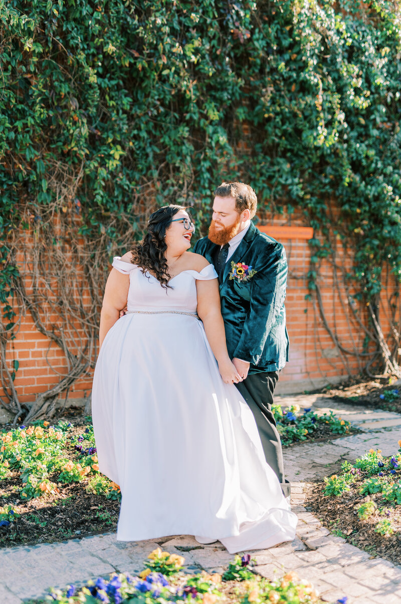 Bride and groom holding hands and smiling at each other while standing outside in front of a brick wall covered with greenery