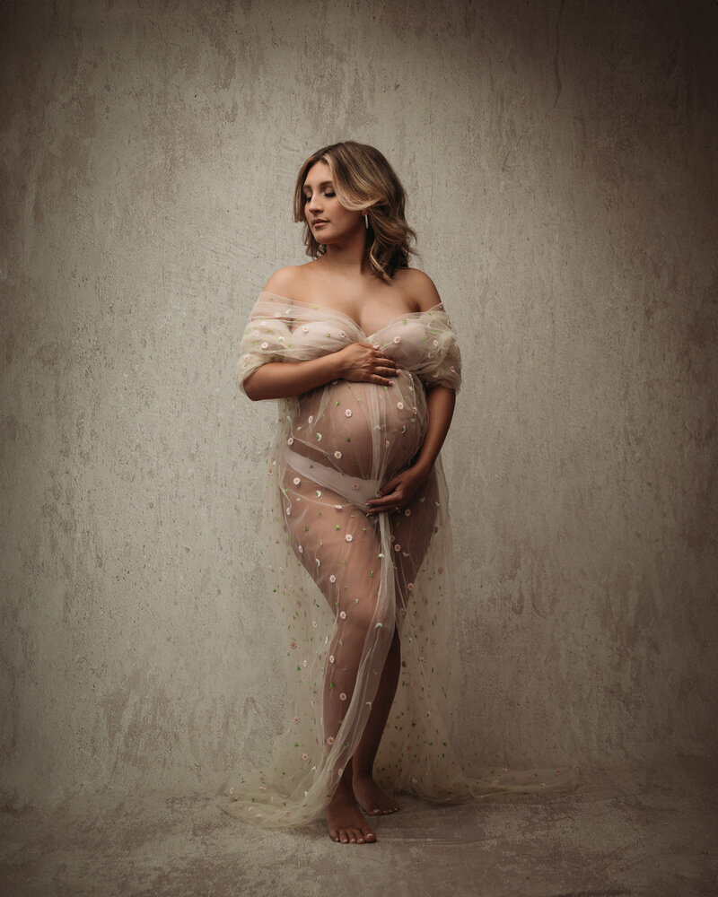 beautiful pregnant woman draped in beige floral sheer fabric in front of cream backdrop
