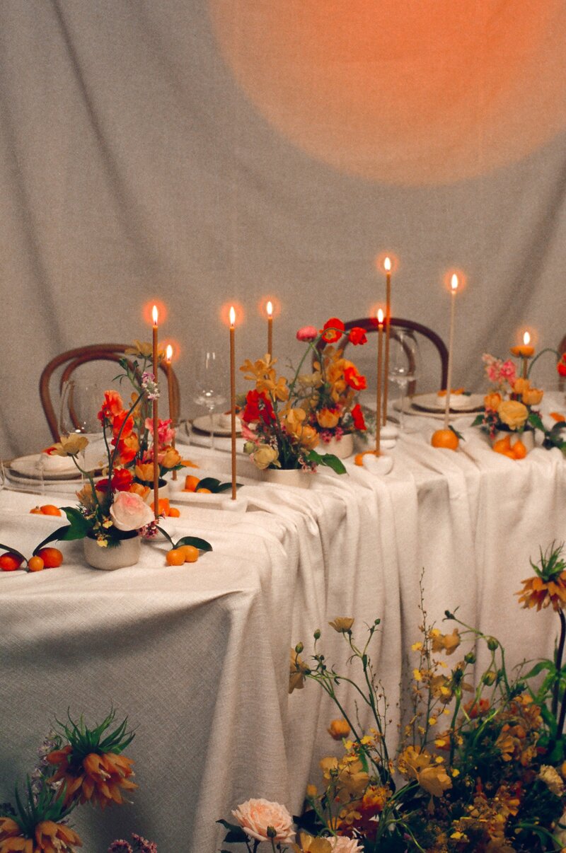 Reception table set with coral and tangerine floral centerpieces, taper candles, and textured linen, styled in an intimate, romantic setting.