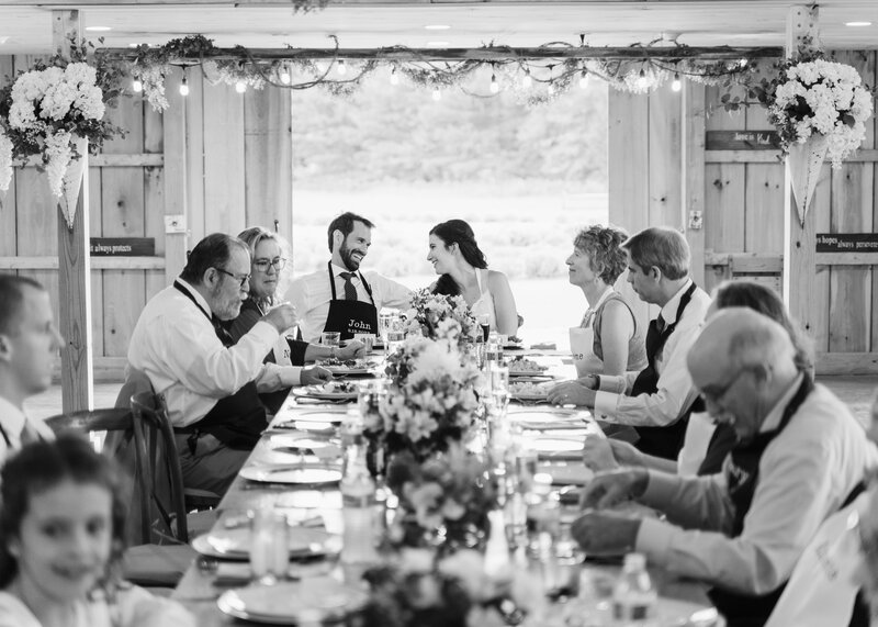 Bride and groom smile at each other at the head of a long farmhouse table surrounded by close family