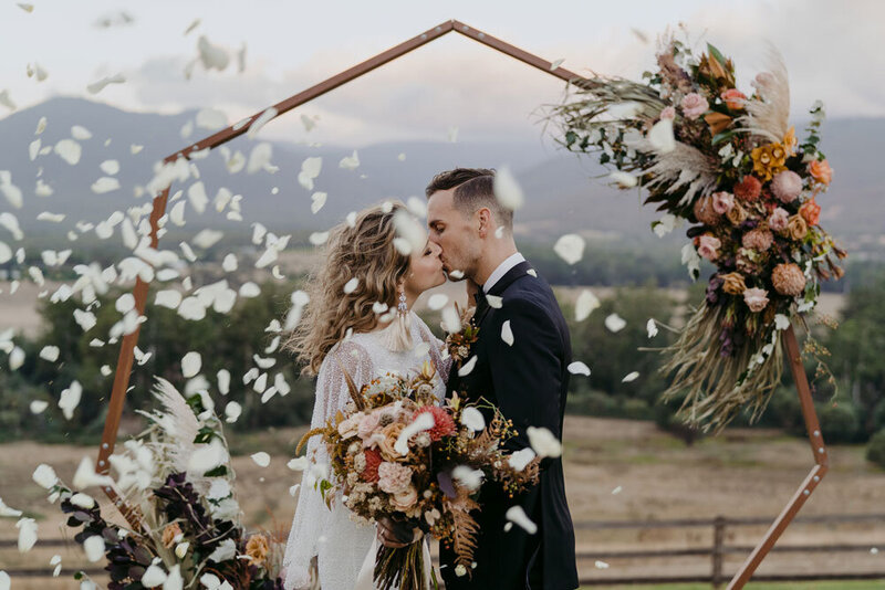 Bride and groom kissing at the alter at the Riverstone Estate, with flower petal confetti and octagon wooden arbour decorated with flowers
