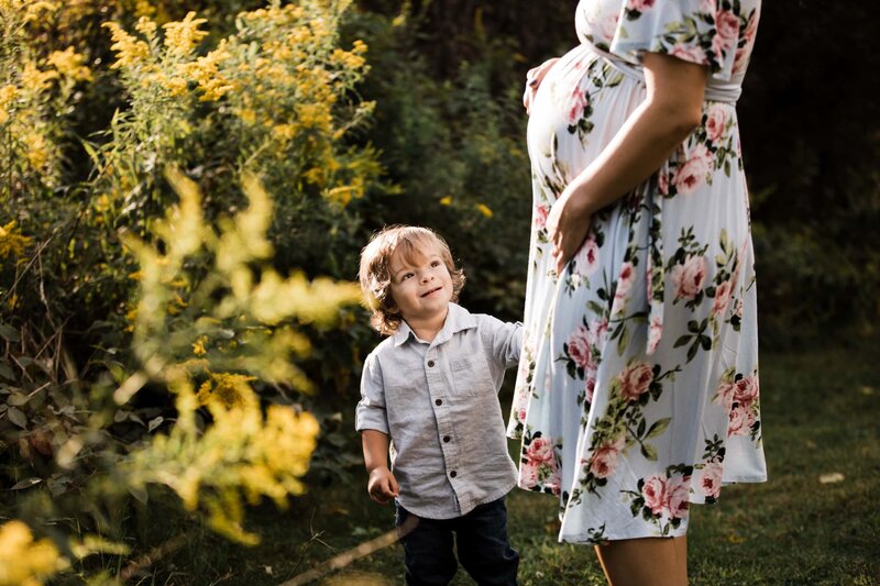 A young boy holding a pregnant woman's hand in a garden with blooming flowers, capturing the moment with a Pittsburgh maternity photographer.