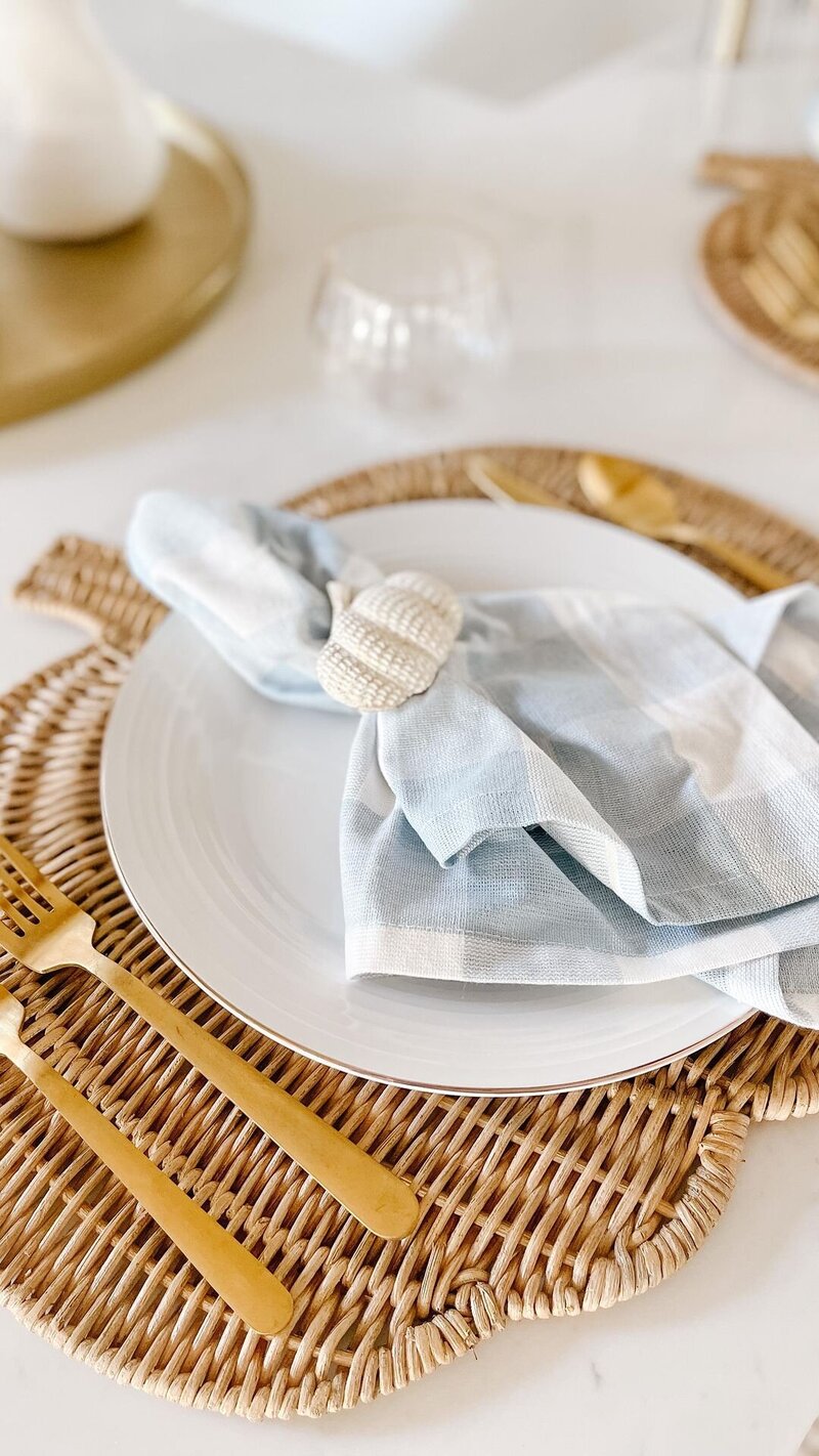 a place setting with gold utensils. a woven place mat, and light blue napkin