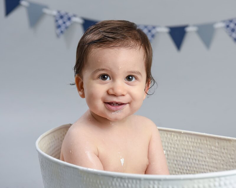 A baby boy sitting in a white tub with wet hair, smiling, and a blue and gray bunting backdrop.
