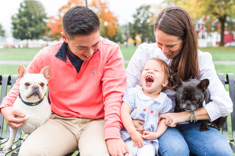 family of three sitting on a bench laughing with two french bulldogs