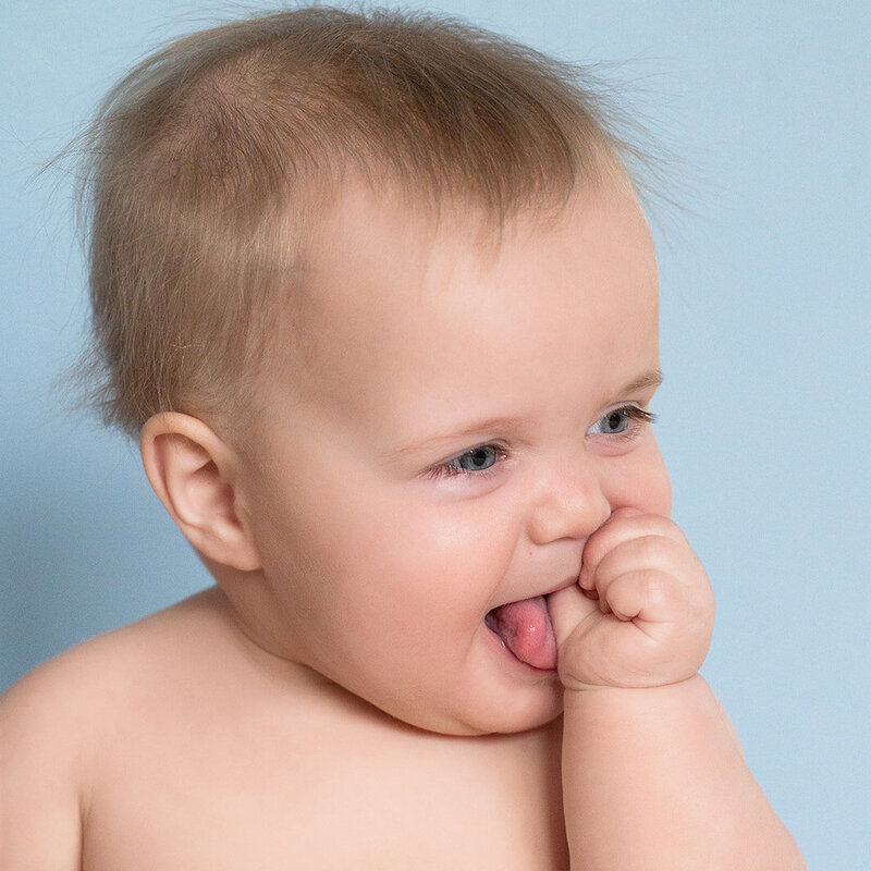 Smiling baby with finger in mouth in front of a soft blue backdrop during a milestone session in Lancaster, PA.