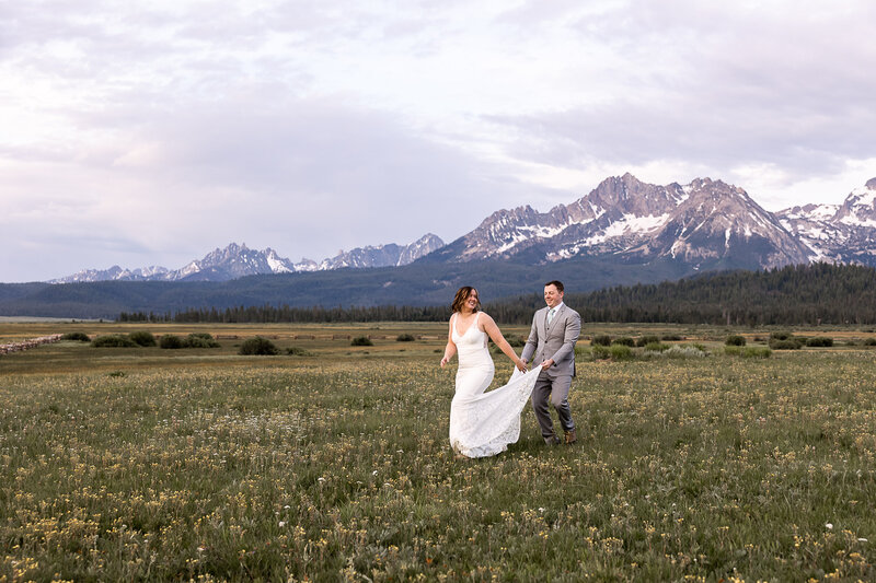 Bride running through a flower meadow with groom running behind holding her dress laughing