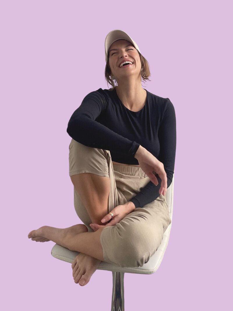 Caitlin sits comfortably on a chair and a joyful smile. She wears a black long-sleeve crop top and beige pants against a blue background.