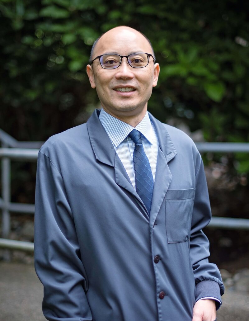 Raymond J. Kao, DMD smiling headshot in dental uniform outside in front of green shrubbery