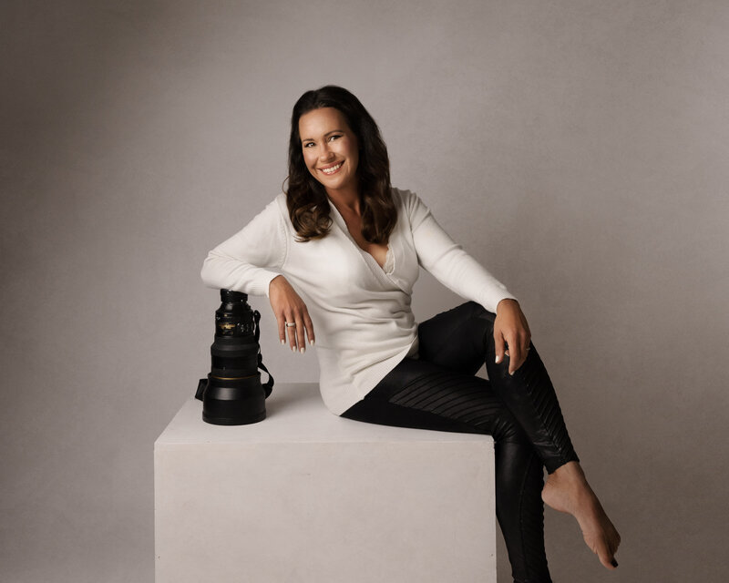 Photo of award winning portrait Artist Kat Figlak.  Woman with brown hair wearing a white sweater with black leather pants sitting resting her elbow on a Nikon 200mm 2.0 lens