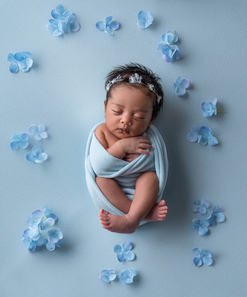Baby girl swaddled in blue and waring a blue headband is sleeping with her feed and hands folded and exposed for newborn photoshoot. She is surrounded by blue flowers. Captured by Bryn Mwar Philadelphia and Main Line photographer Liza Nicole.