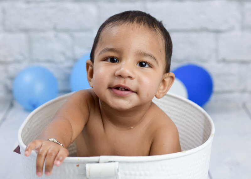 A baby boy sitting in a white tub surrounded by blue and white balloons, smiling with a gray brick wall background.