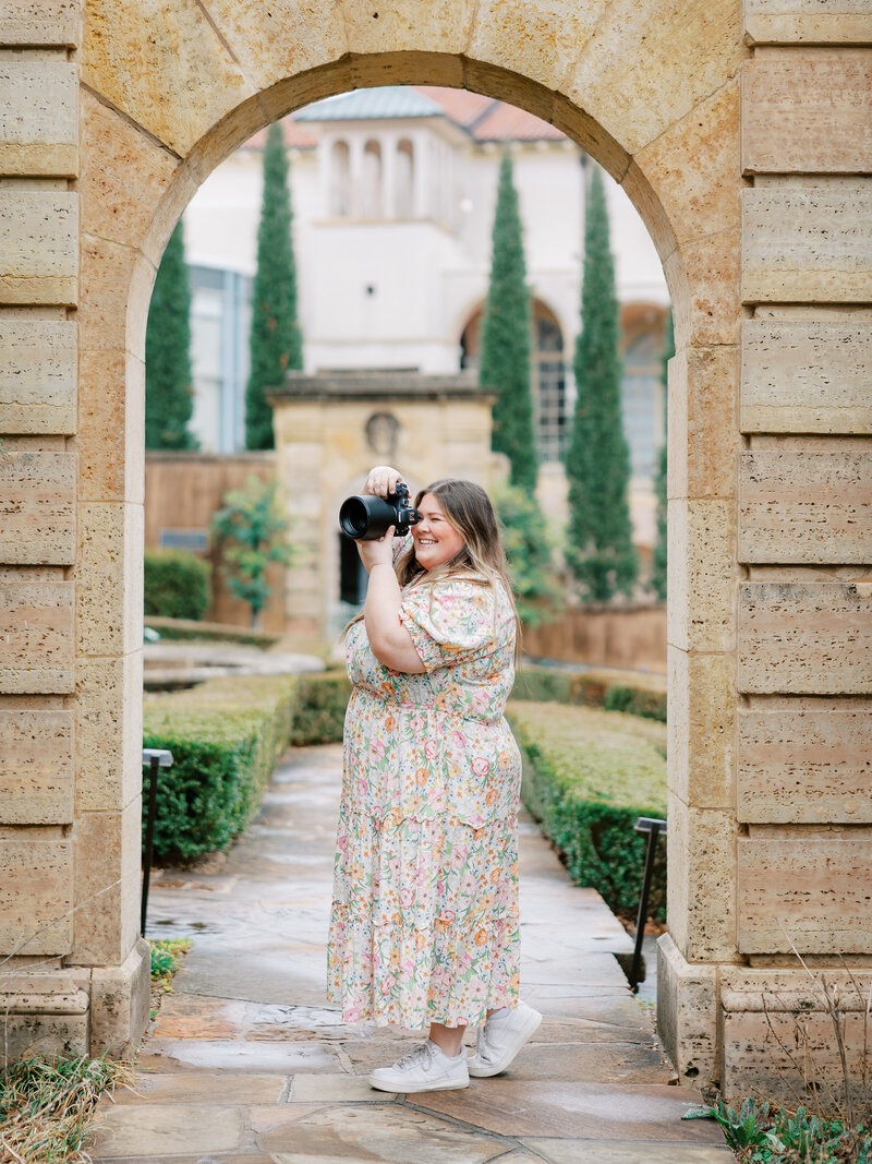 Shalae stands in floral dress taking a picture on her camera under archway at Philbrook Museum in Tulsa, Oklahoma