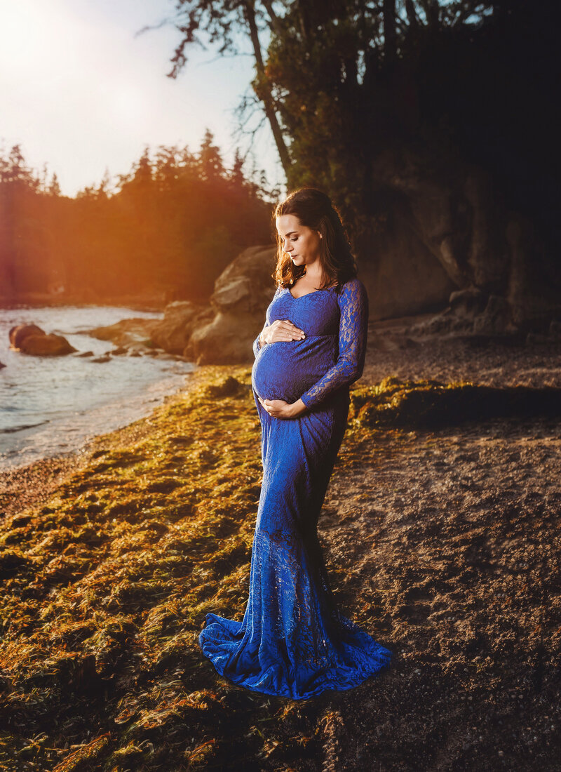 Beautiful pregnant woman poses for photo and embraces her belly in a full length blue gown in nature