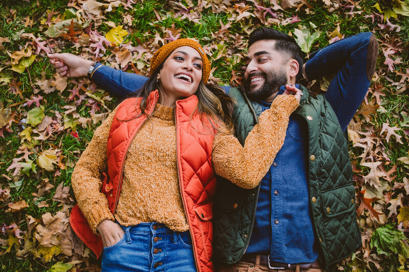 Couple sharing a laugh together in autumn clothing laying on the grass with fall leaves all over
