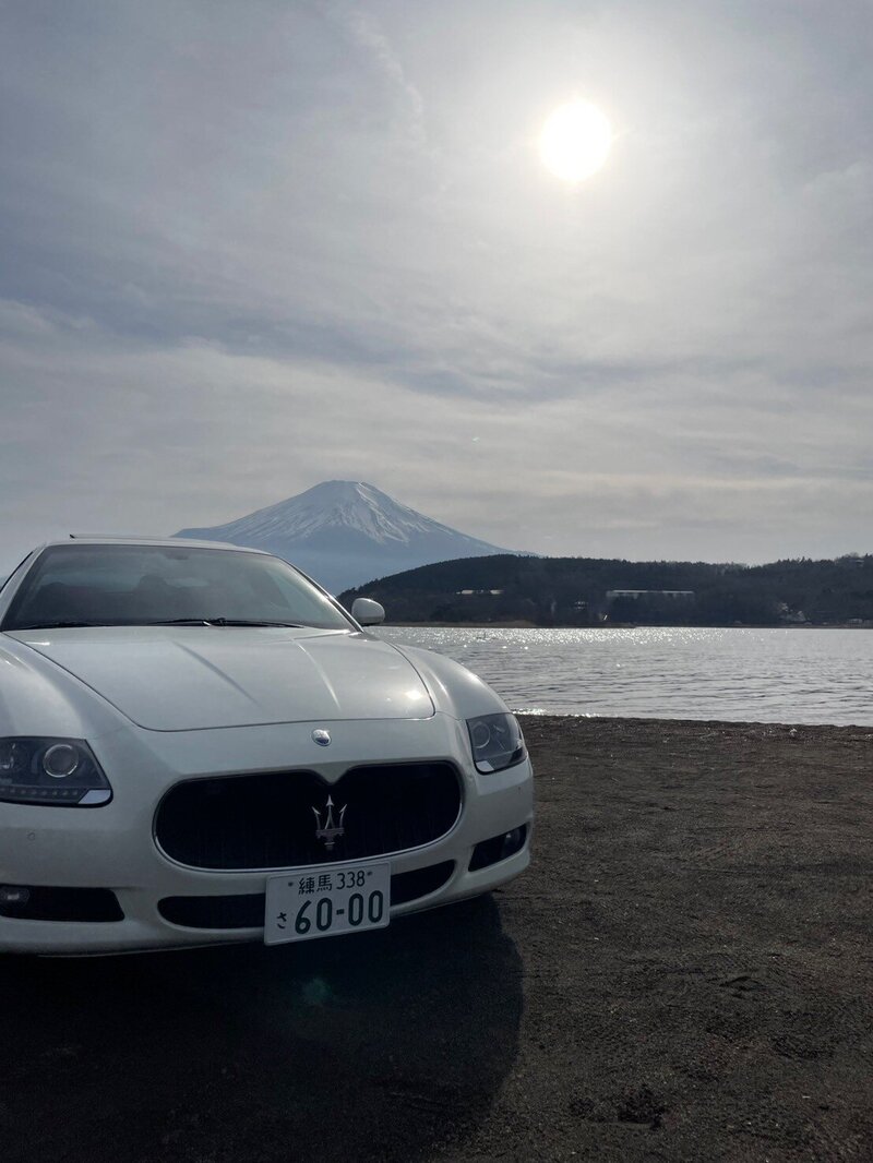 Front view of a white Maserati parked lakeside with the majestic Mount Fuji and the sun overhead in the background, creating a serene and luxurious scene.
