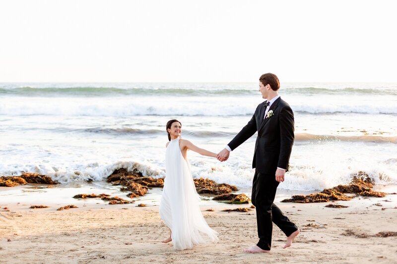 fashion forward bride walks with groom across newport beach