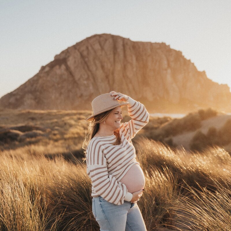 Pregnant Mother holding her belly and her hat while posing in front of the rock in Morro Bay.