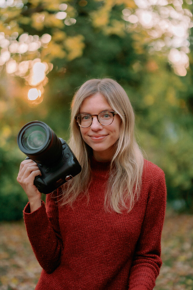Nikki Robinson holds a camera outdoors while wearing a red sweater.