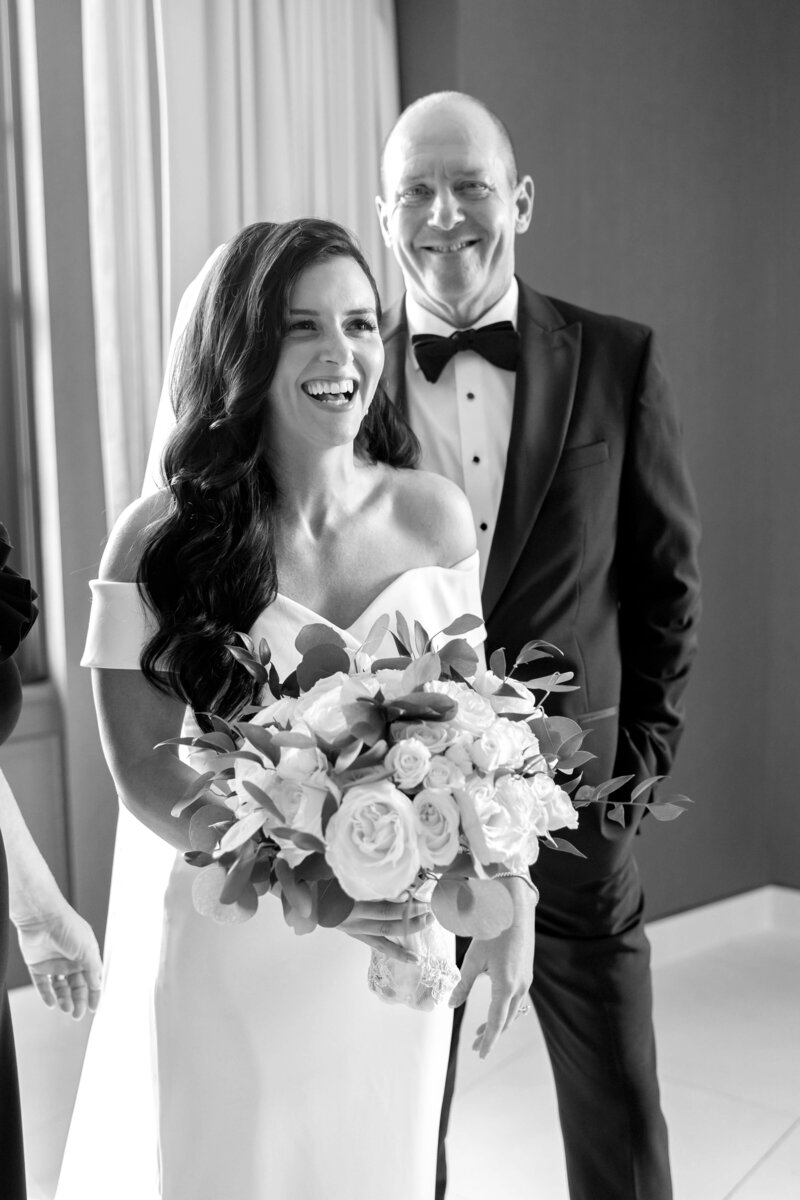 A smiling bride in a white dress holds a bouquet of white and pink flowers, standing next to a man in a tuxedo. They are indoors, with a curtain in the background. The image is in black and white.