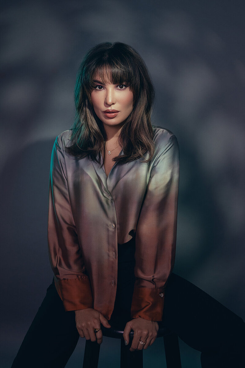 Moody studio portrait of Lydia Bell wearing a satin blouse, seated under dramatic lighting with patterned shadows, creating a cinematic and introspective vibe.