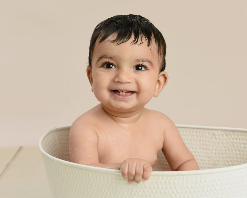 A smiling baby with spiky hair sitting in a white tub, surrounded by neutral  decor in a beige background.